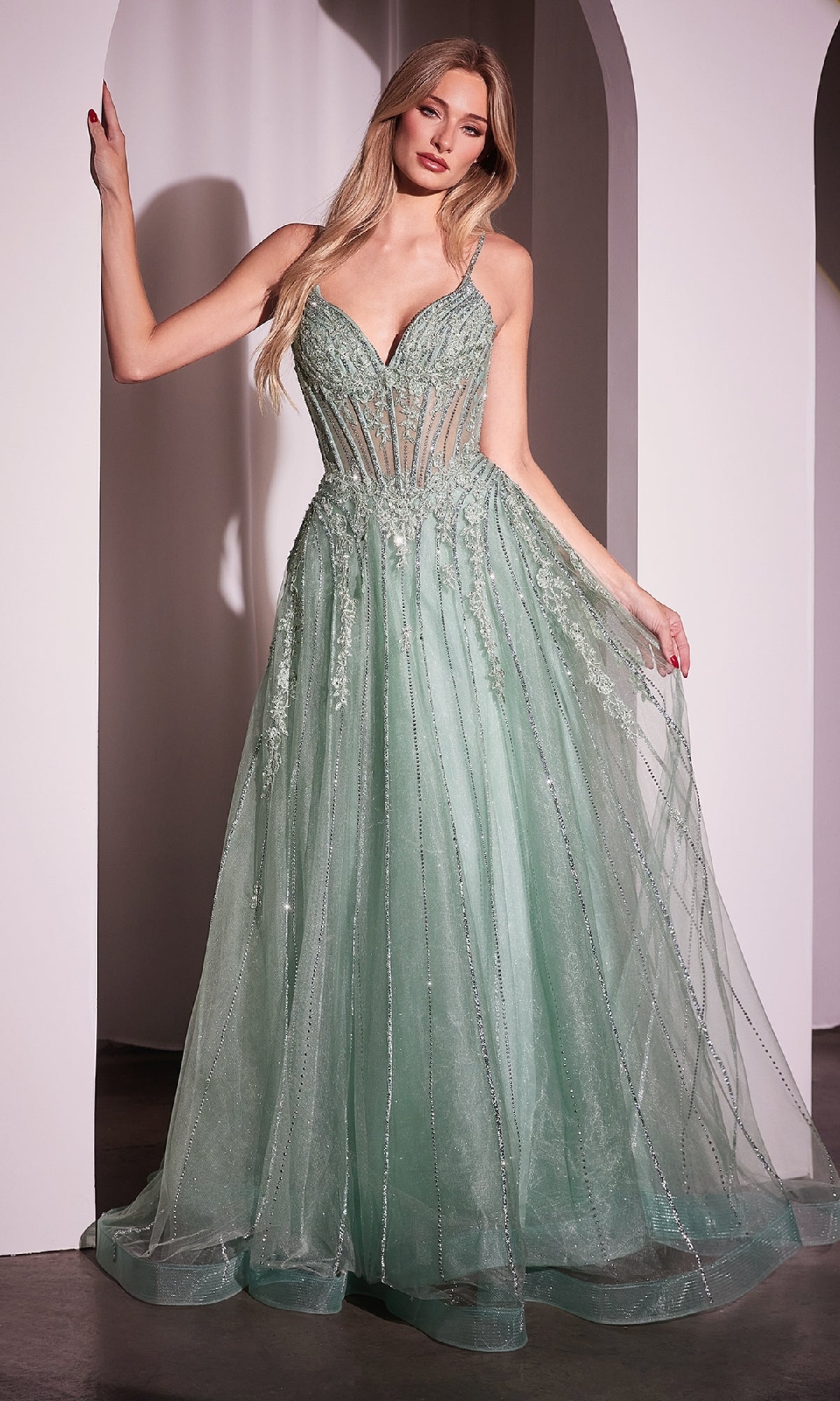 A woman stands indoors wearing the Ladivine CD786 Crystal-Beaded Long Prom Dress, a mint green, floor-length gown with a sheer bodice, thin straps, and sparkling embellished details.